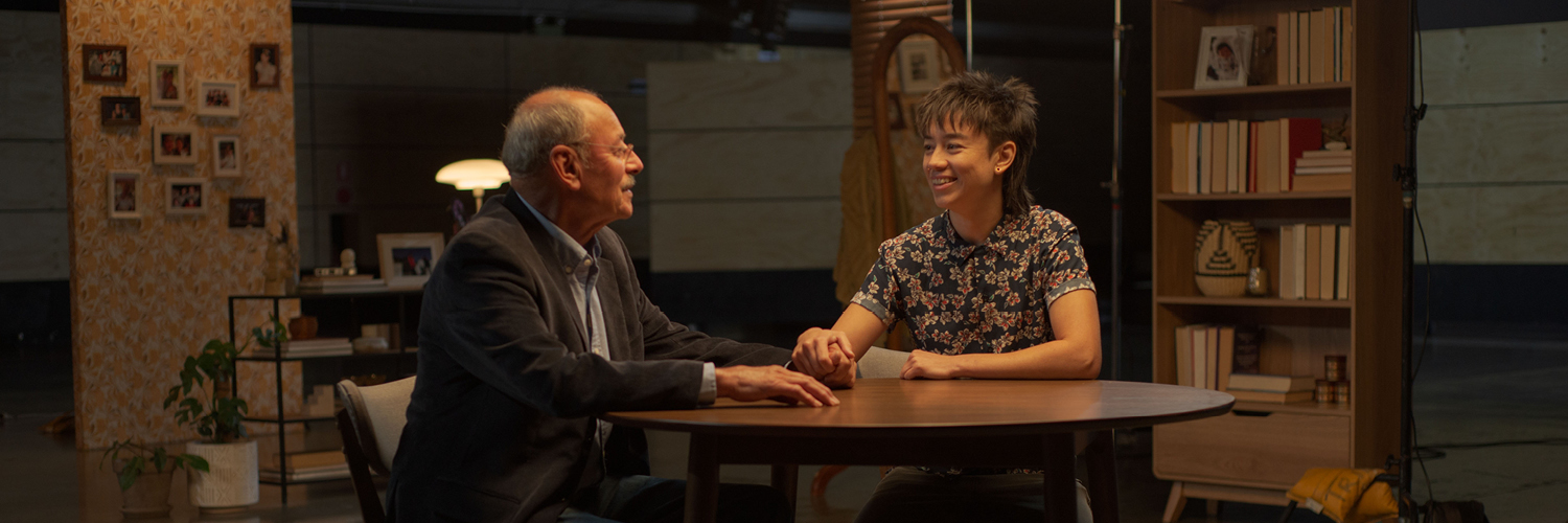 An older father and his adult child hold hands and smile at each other, sitting at a table 