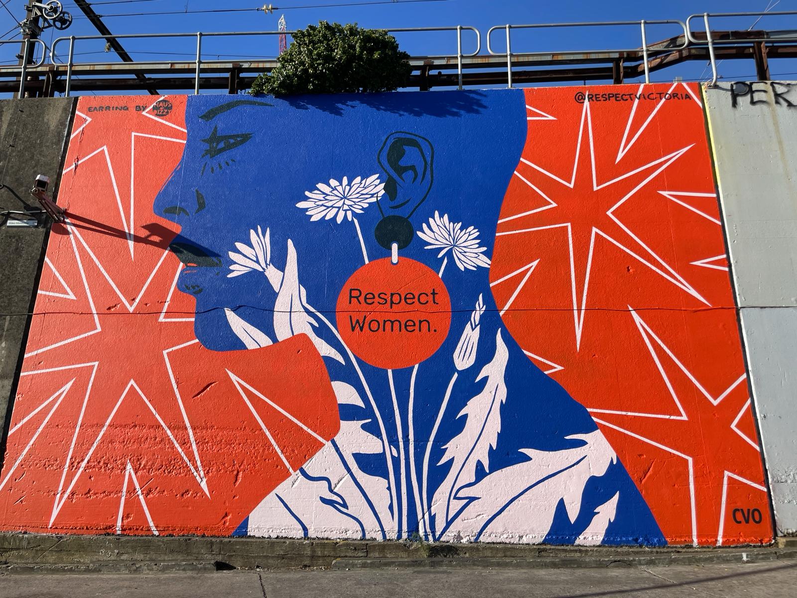 A large mural showing the blue silhouette of a woman's head and shoulders. She wears orange statement earrings that read "Respect Women.". White dandelions cover her shoulder and neck. There are white starbursts on the orange background. 