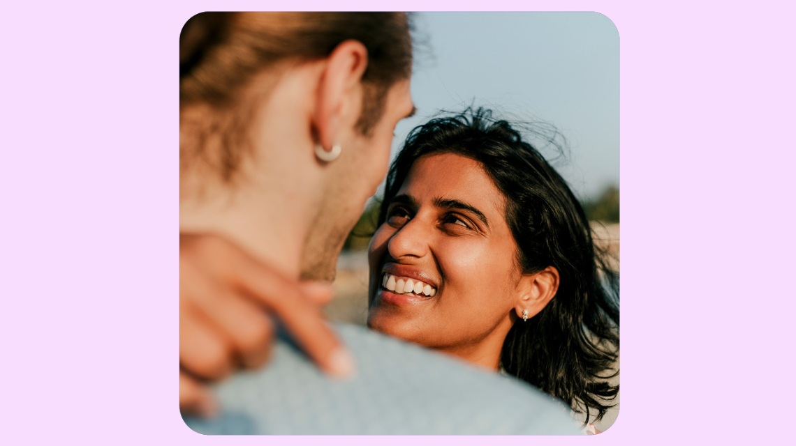 A close up of a couple hugging, we see the woman's smiling face and her partner's shoulders. 