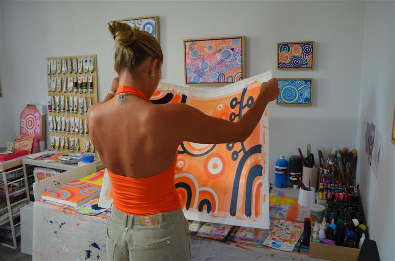 Bayley Mifsud working in her studio. Her back is to the camera and she lifts the canvas to see the incomplete artwork.